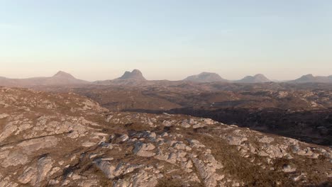 Aerial-view-flying-over-a-remote-rocky-area-of-Assynt-towards-the-famous-mountains-of-Canisp,-Suilven,-Cùl-Mòr-and-Stac-Pollaidh-on-a-sunny-spring-evening