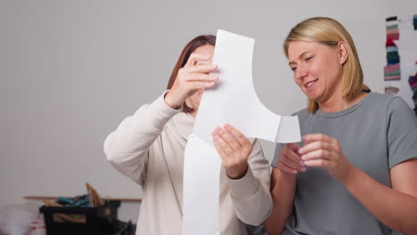 two female tailors smiling as they review fabric cutout pattern during discussion, surrounded by sewing materials, fabric samples, and colorful textile swatches in creative bright workspace