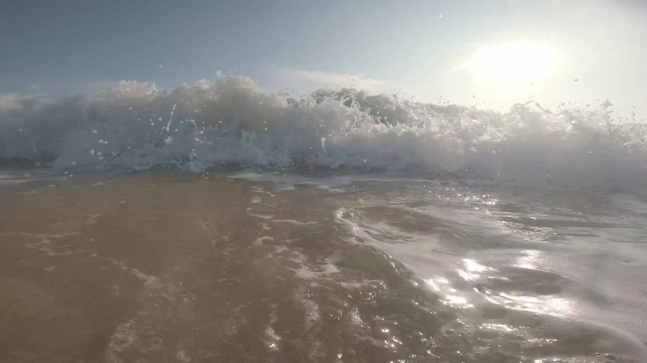 A low water-level view captures foamy surf receding before an Indian Ocean swell rolls toward the lens and breaks in bright sunlight at Mirissa Beach, Sri Lanka.