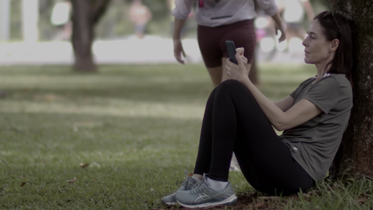 Beneath trees where shadows spark, seated with phone in the local park