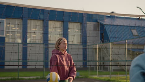 vista trasera de la dama con capucha azul sirviendo voleibol a la dama con hoodie marrón, que lo sostiene con expresión seria, otra dama se extiende mientras la pelota cae en el suelo, el fondo presenta un edificio