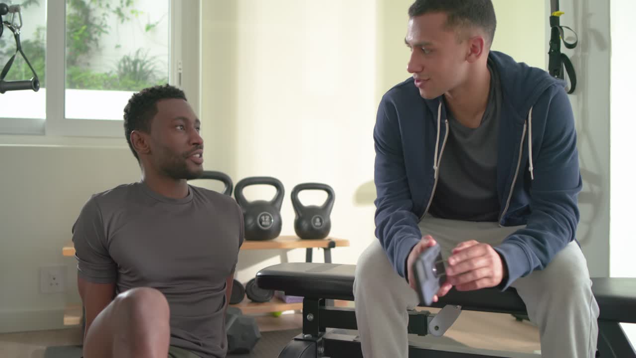Compañeros masculinos diversos tocando un smartphone, compartiendo consejos de fitness y bebiendo agua junto a un banco en un gimnasio en casa.