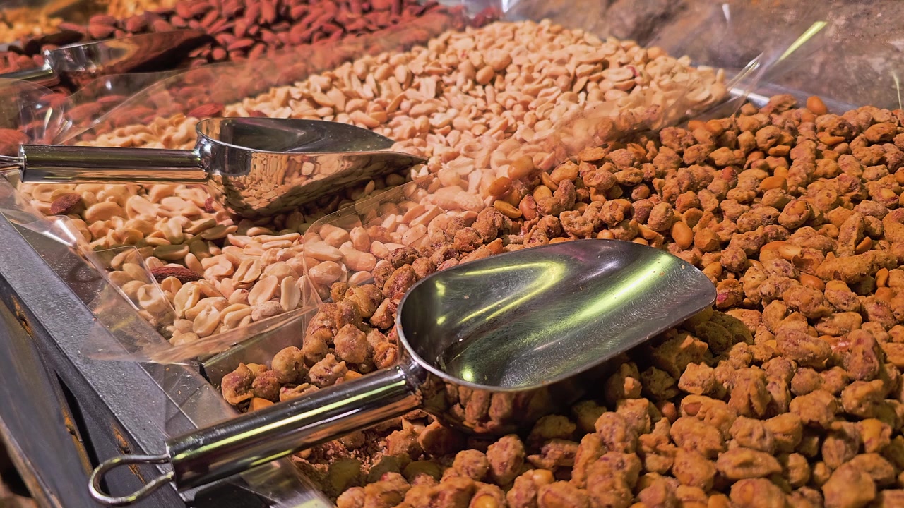 Detailed view of assorted nuts in clear bulk bins, showing cashews, peanuts, almonds, and coated snack mix with metal scoops, warm indoor light, and self serve market display.