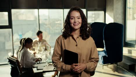 Happy-brown-haired-girl-with-wavy-hair-in-brown-business-clothes-looks-at-the-camera-and-smiles-in-a-large-office