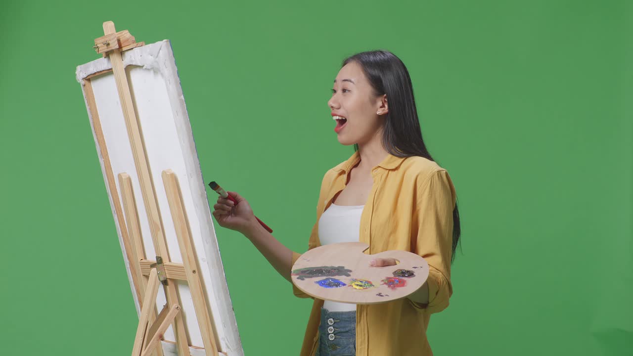 아시아 여성 예술가가 캔버스 위의 그림을 보고 와우라고 말하면서 녹색 스크린 스튜디오에서 오일 페인트와 브러쉬로 그림을 그리고 있습니다.