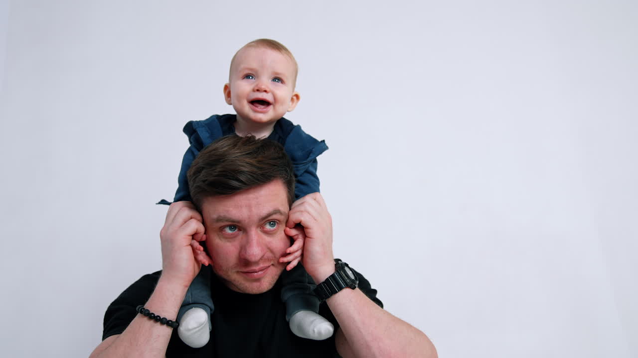 Happy smiling infant boy sitting on dad's shoulders. Father and son concept. White backdrop.