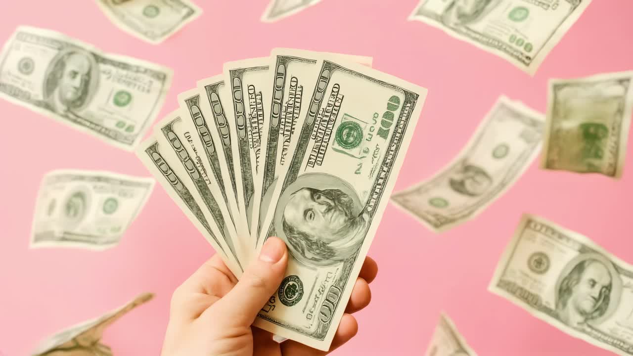 A hand holding dollar bills with more floating around against a pink background Captured from a low