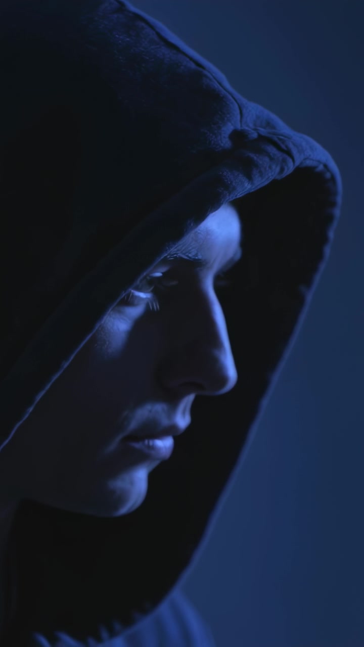 Video verticale: Uomo in felpa con cappuccio, che reagisce a una luce di contorno blu, alzando la testa per rivelare il profilo in studio.