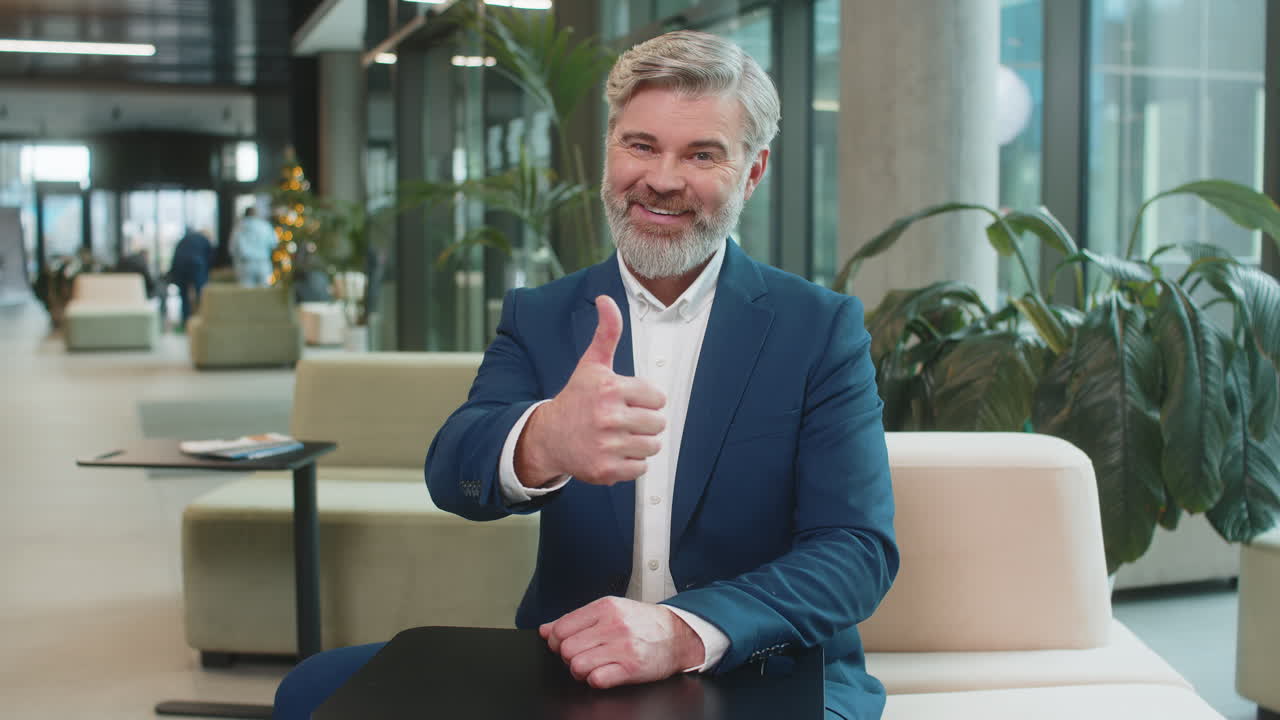 Happy mature caucasian businessman in suit looking at camera showing thumb up in modern office lobby
