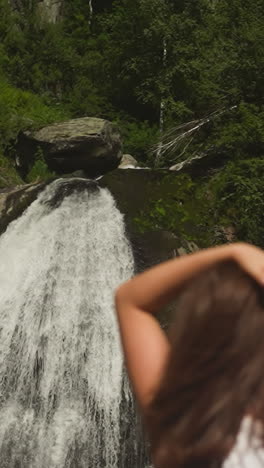 mother adjusts long hair holding toddler son rest by waterfall in rocky park slow motion. young woman shows with boy amazed by water cascade in wood