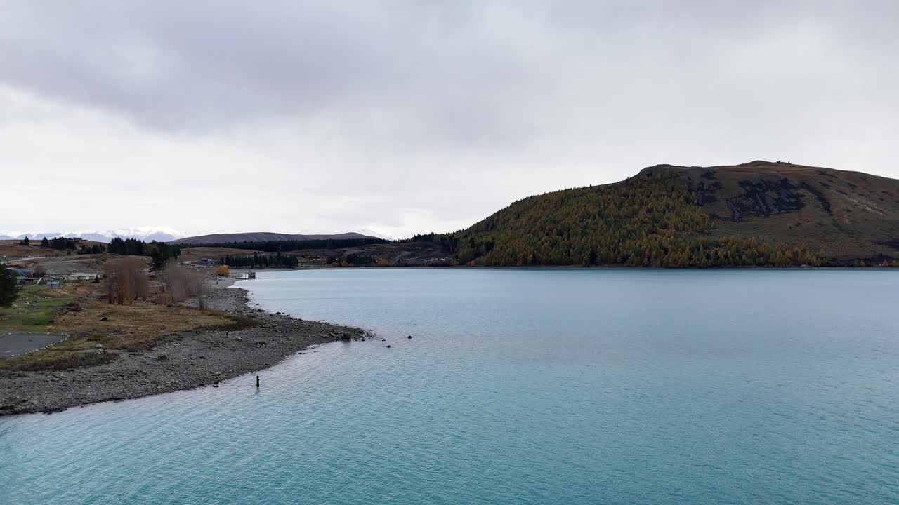 A tranquil view of Lake Tekapo with calm waters and surrounding hills under overcast skies, captured in a steady aerial shot