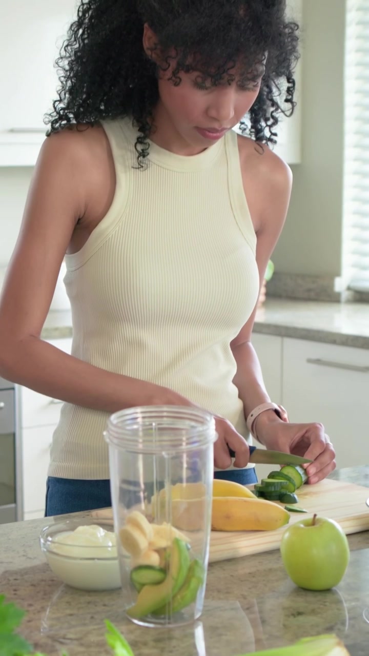 Video verticale: Donna con top a costine versa liquido in una tazza, taglia un cetriolo in cucina mentre prepara una bevanda.
