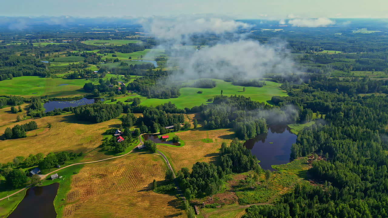 Aerial overview orbit around misty thin clouds with sunlight cascading down on forest landscape with fields and ponds below, beautiful, serene scene