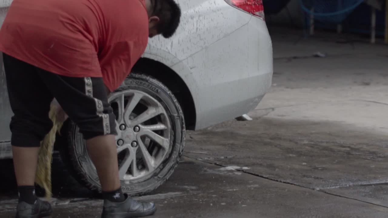 Person washing a car wheel with a sponge in a wet, outdoor setting during daytime