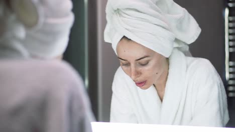 Eine-Frau-Mit-Einem-Handtuch-Auf-Dem-Kopf-Wäscht-Ihr-Gesicht-Mit-Wasser-Und-Blickt-In-Den-Spiegel
