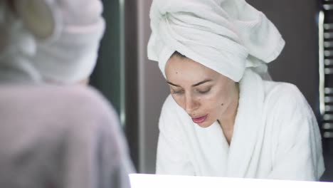 a woman with towel on head washes her face with water, looking at the mirror