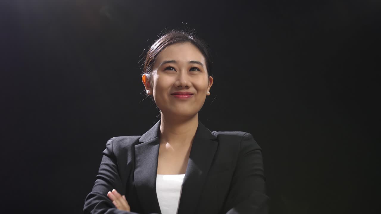 close up de una mujer sonriente de habla asiática en traje de negocios cruzando los brazos mientras está de pie en el estudio de pantalla negra