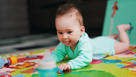 beautiful baby boy lying on the colorful mat clapping hands by the floor. funny kid stuck out his tongue and looks at the bottle in front of him.