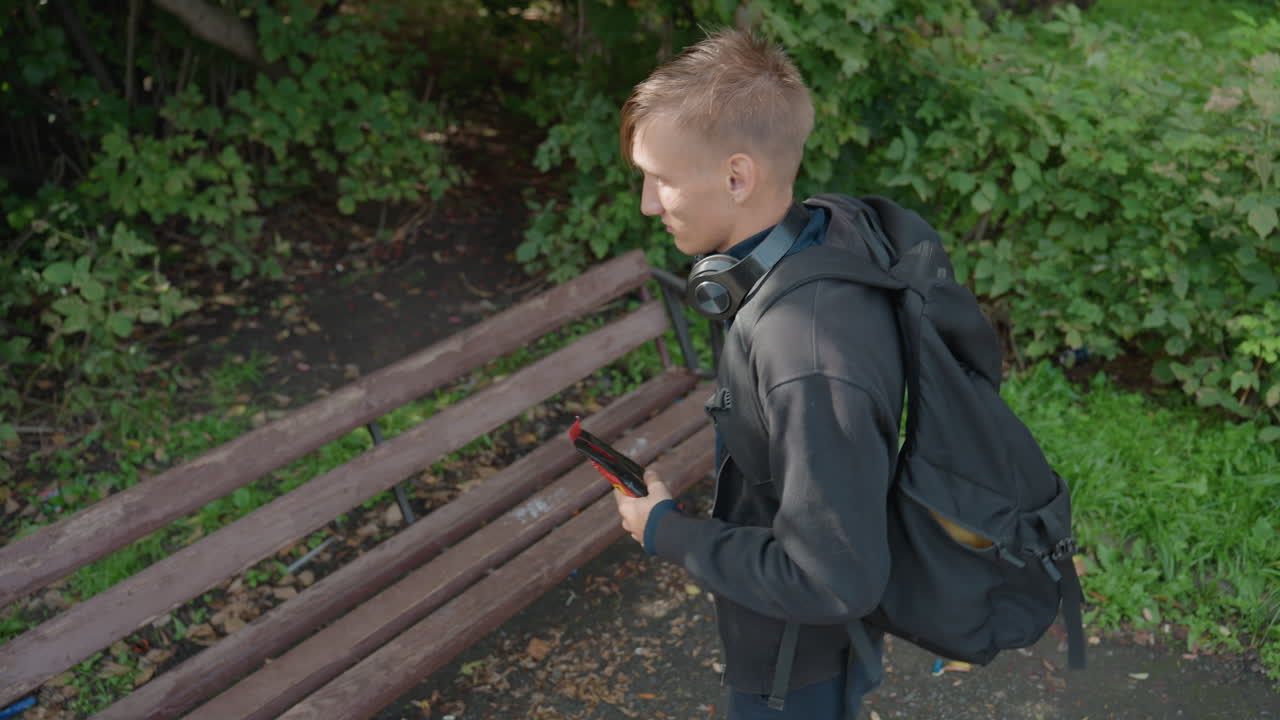 Young man examines wrapper on park bench, Gentle daylight highlights young man inspecting snack package outside, Casual young man seated on bench inspects snack wrapper in leafy park environment