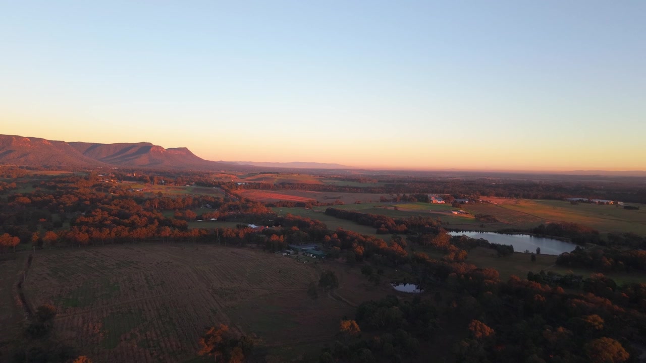 Aerial sunrise across Hunter Valley plains with mountains and lake, drone dolly with soft yellow glow on horizon, countryside of Newcastle NSW Australia