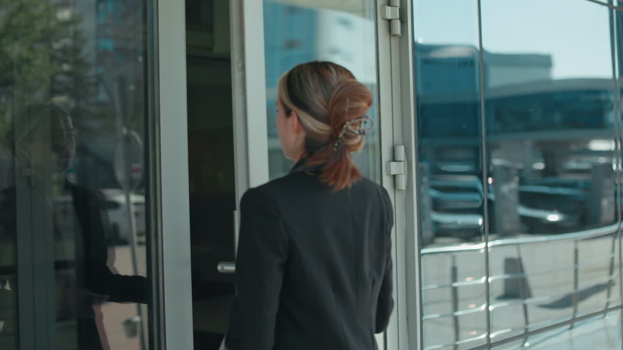 Business woman enters work building with close-up of glass door handle and reflections of busy urban street, parked cars, and passersby visible on glass surface