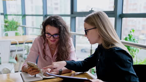 two young women look at smartphone and discuss something smiling. female office workers talk about ideas.