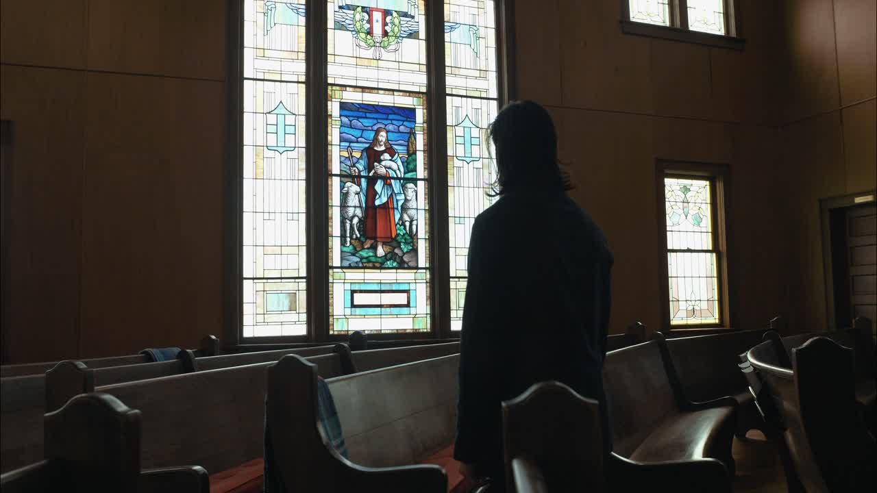 A lone figure stands in the pews of a church, facing a stained-glass window, symbolizing prayer, contemplation, and the search for meaning