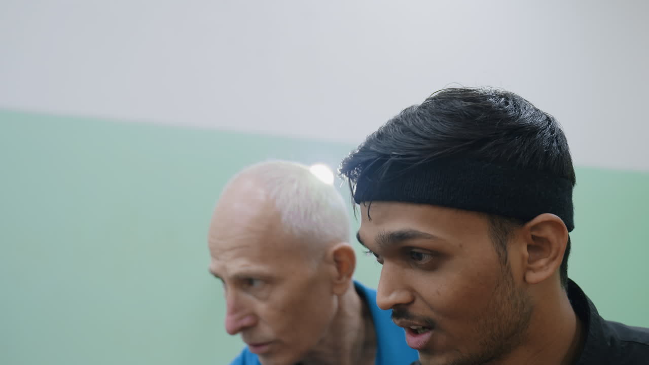 Elderly man guiding young adult on maintaining stable posture during coaching session, demonstrating proper stance and control while teaching balance with light shining in indoor sports training hall