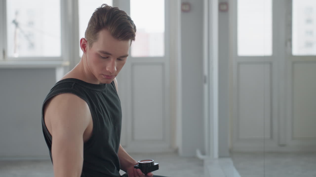teenager seated facing large mirror, suitcase and makeup kit next to him on floor, reaches for black makeup container, opens lid, inspects reflection under soft daylight through windows in studio