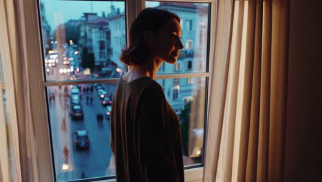 woman looking out apartment window at city street at dusk