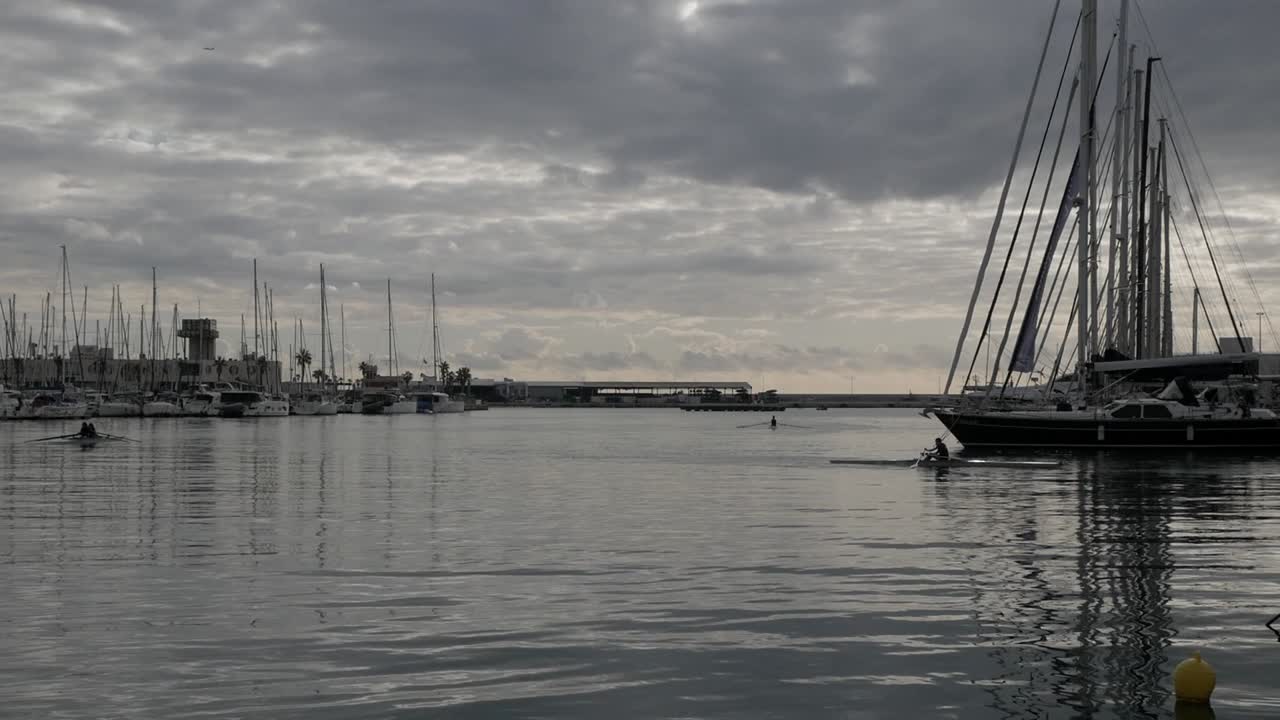 A serene harbor with rowers and sailboats under a cloudy horizon in Alicante, Spain