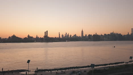 AERIAL:-Rising-up-over-East-River-overlooking-Manhattan-New-York-City-Skyline-in-Beautiful-Dawn-Sunset-Orange-Light