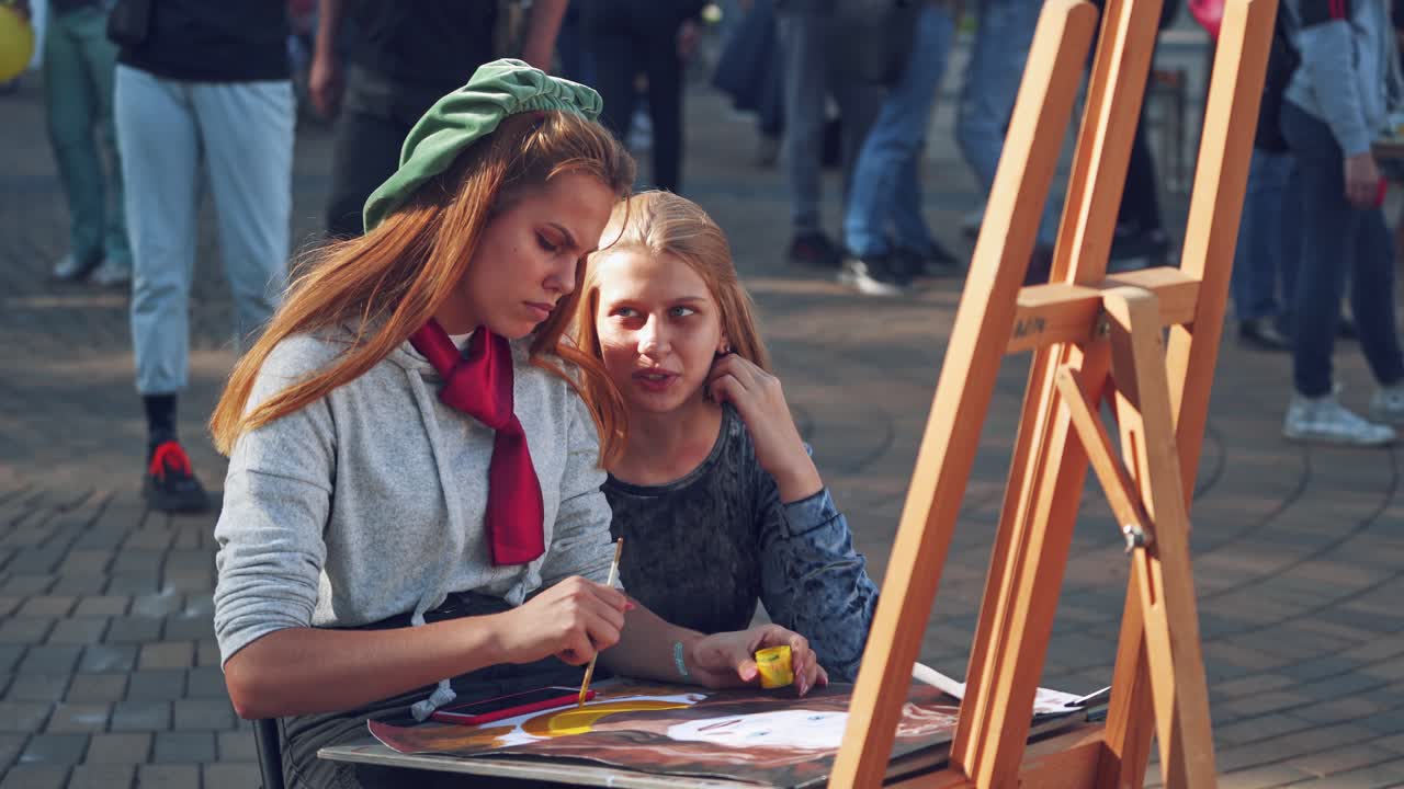 Two women near easel. Beautiful girl artist painting with brush on the busy street background. Female friend talking to female artist outdoors.