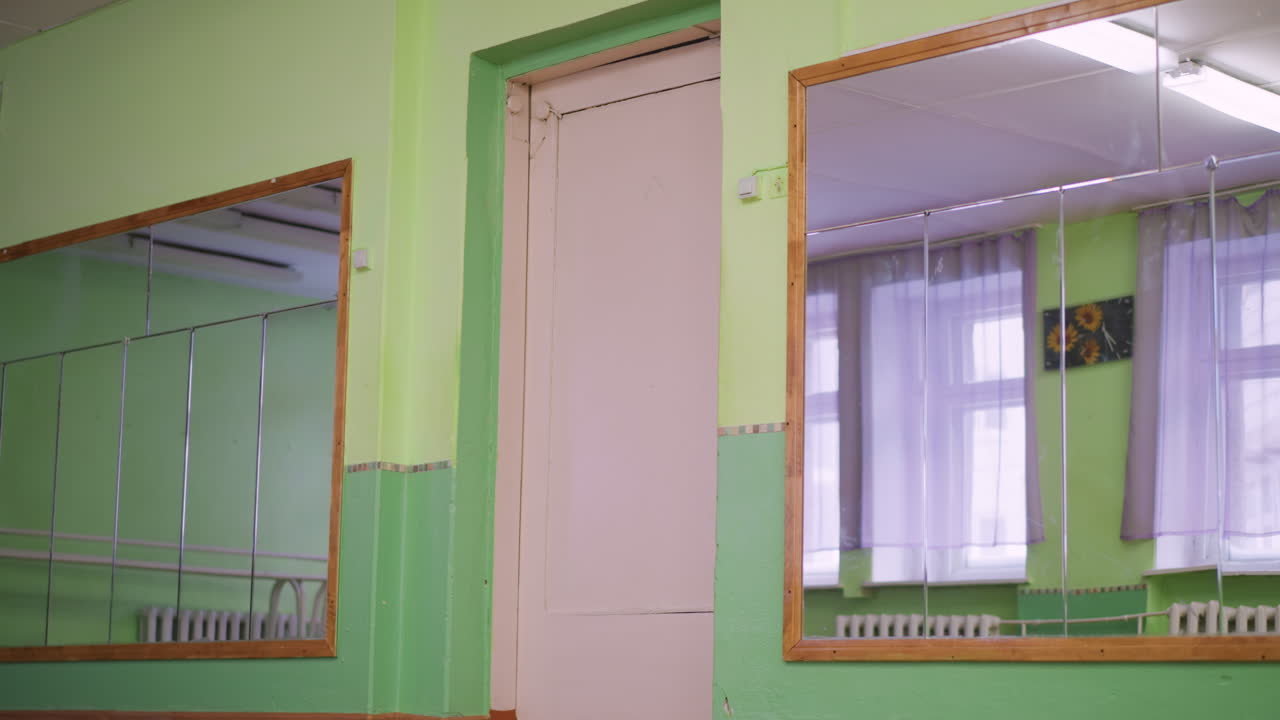 Green interior with two wall mirrors and open door, reflections of windows and radiators over wooden floor, bright studio mood hinting recent exit by lady who walked out calmly