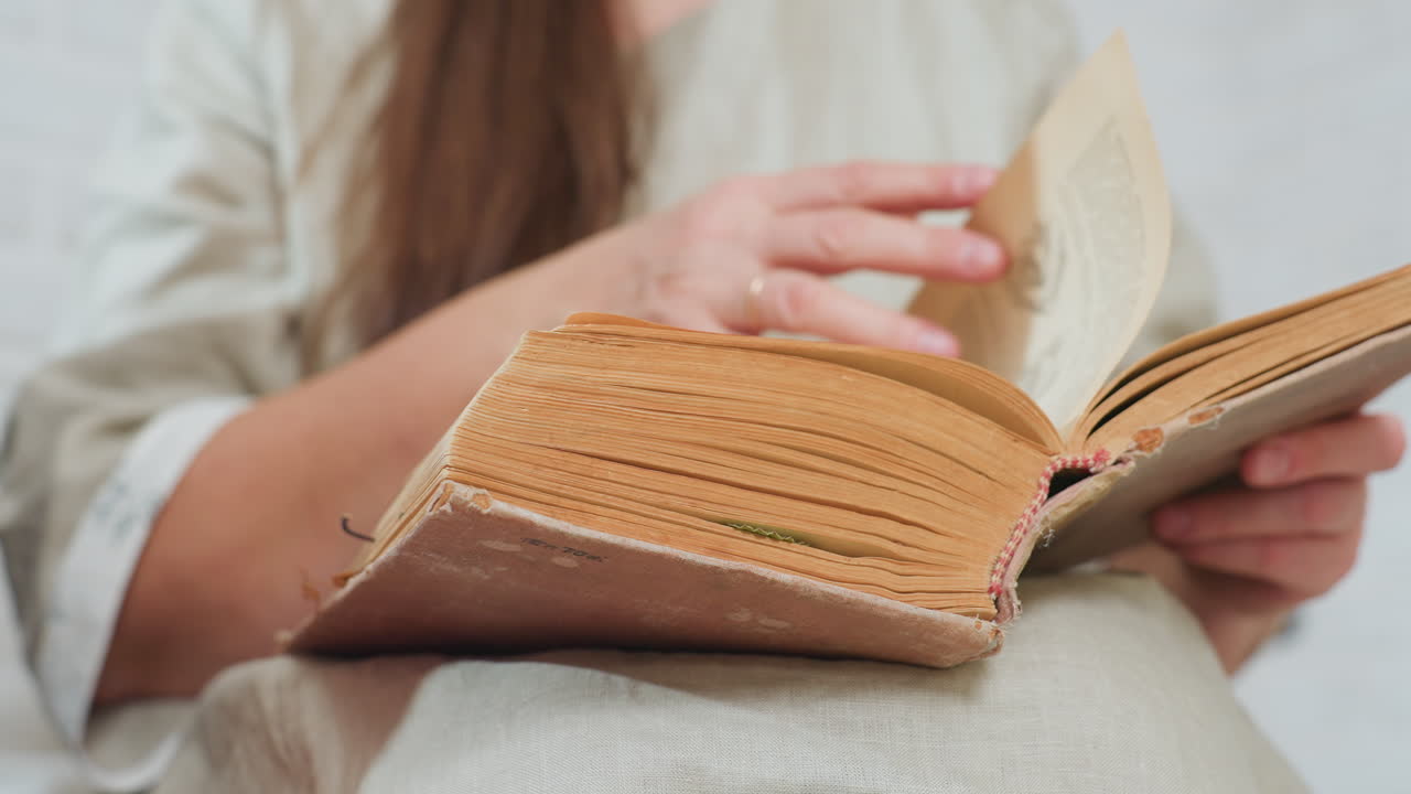 partial view of aged woman in beige clothing flipping pages of worn old book, fingers gently turning fragile paper, texture of aged pages visible, natural indoor light creating warm
