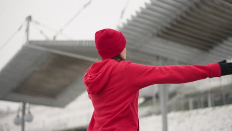 vista trasera de una mujer con guantes estirando los brazos durante el entrenamiento al aire libre bajo un techo de metal con colina nevada y postes de luz en el fondo, mostrando enfoque y energía en un ambiente frío de invierno