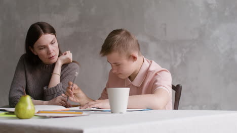 boy with down syndrome drinking from a cup and doing homeworks sitting at table in the living room at home. his mother helps him