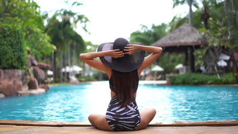 A-woman-sitting-crossed-legged-on-the-edge-of-a-swimming-pool,-in-a-bathing-suit-with-her-back-to-the-camera,-raises-her-arms-to-adjust-her-sun-hat