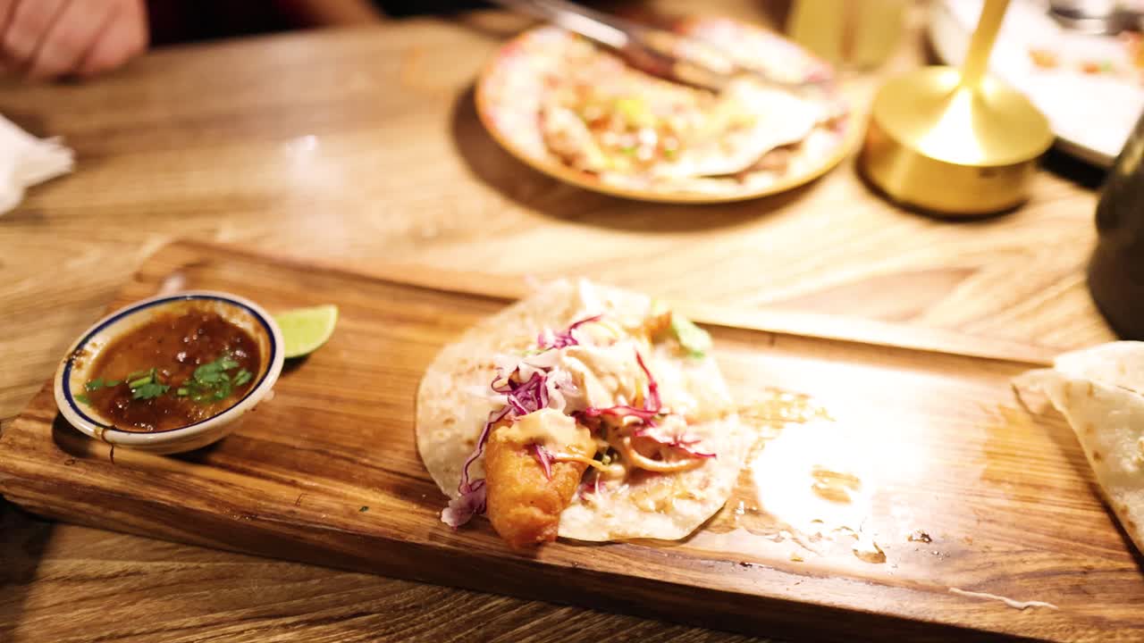 En hånd presser lime over tacos på et træbræt, hvilket fremhæver den farverige ret under varm, stemningsfuld belysning.