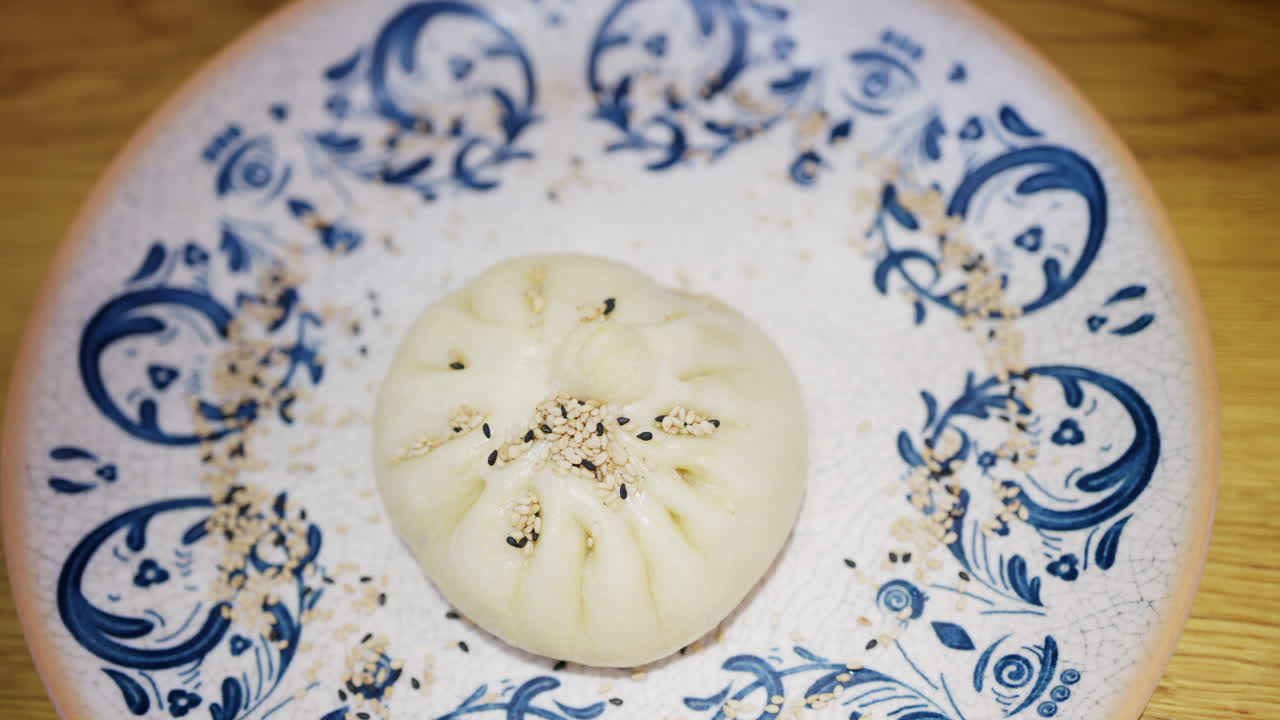 Primo piano di un panino al vapore guarnito con semi di sesamo su un piatto di ceramica decorativo.