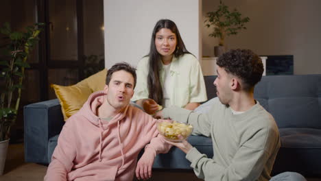 Tres-Amigos-Viendo-Una-Película-Cómica-En-La-Televisión-Sentados-En-El-Sofá-Y-En-El-Suelo-Y-Comiendo-Palomitas-De-Maíz-Y-Papas-Fritas