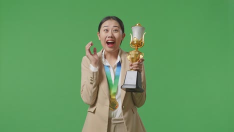 asian business woman in a suit with a gold medal looking at a gold trophy in her hands and saying wow being happy winning as the first winner on green screen background in the studio