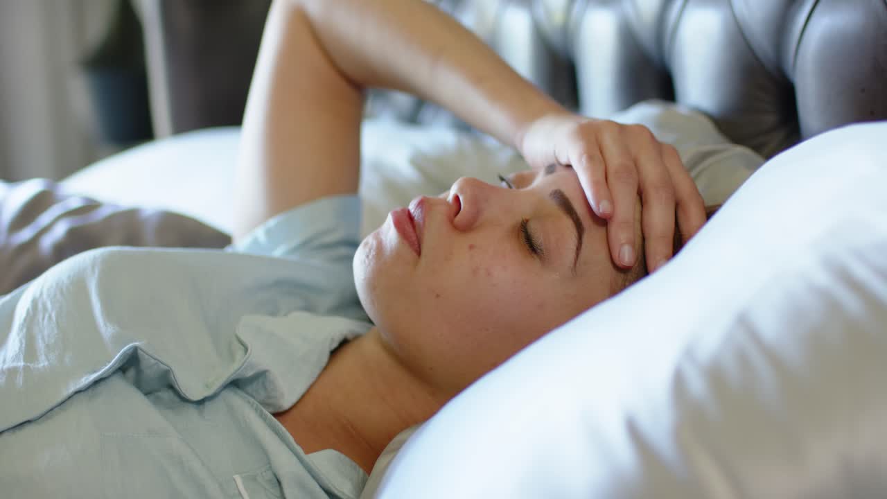 Mulher afro-americana adulta na cama, se revirando e franzindo a testa, levando a mão à testa para aliviar a dor de cabeça.