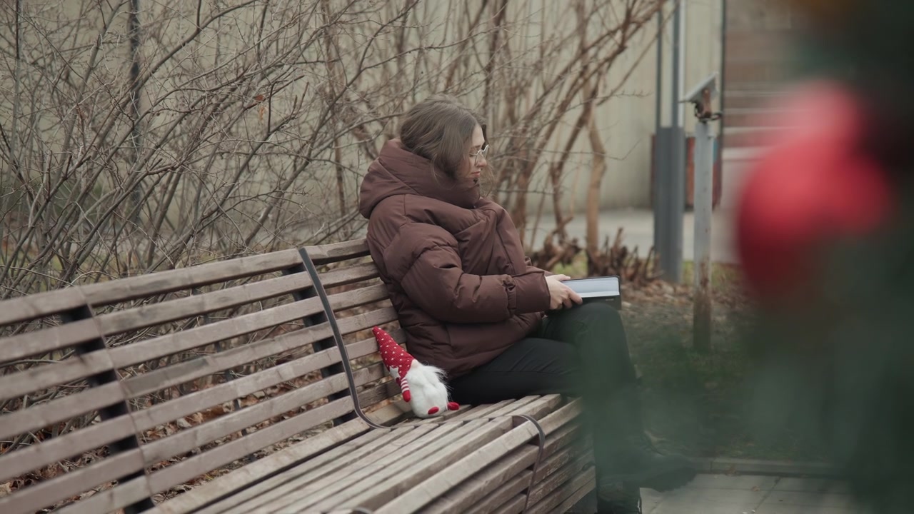 Caucasian woman on bench sketching gnome, blurred red blossom foreground, brown coat and focused profile, intimate courtyard setting with soft bokeh and calm mood, creative pause captured candidly
