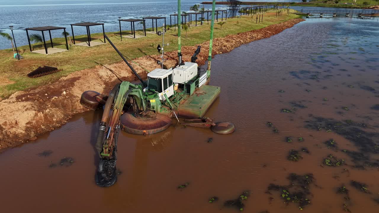 Amphibious dredging machine working in shallow, muddy river water near the shore, cutting into the sediment with hydraulic arms, Posadas, Misiones, Argentina, aerial orbit