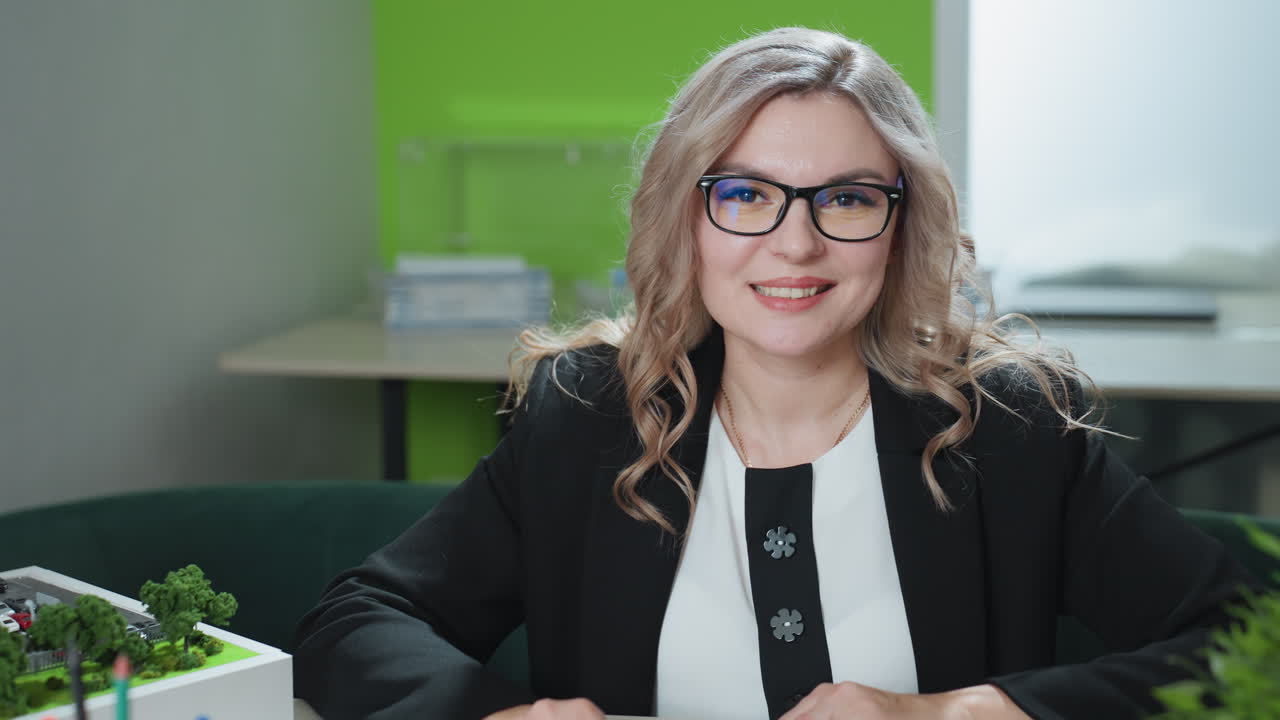 confident woman wearing glasses sits at desk with slight smile, working on laptop in modern office environment with architectural model, green wall, desk supplies, and potted plant