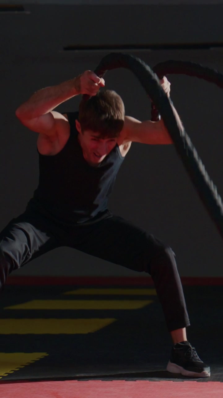 En mann trener intenst med battleropes i et treningsstudio. Han fokuserer med viljestyrke, og musklene anstrenger seg under det svarte utstyret. Svette markerer innsatsen hans på en rød og gul matte. Flott for trening og treningsøkter.