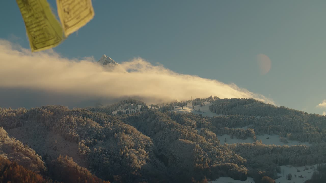 A handheld shot of a snowy mountain peak rising above fog with Tibetan prayer flags in the foreground. The calm daylight scene shows the mountain lit by soft winter sunlight