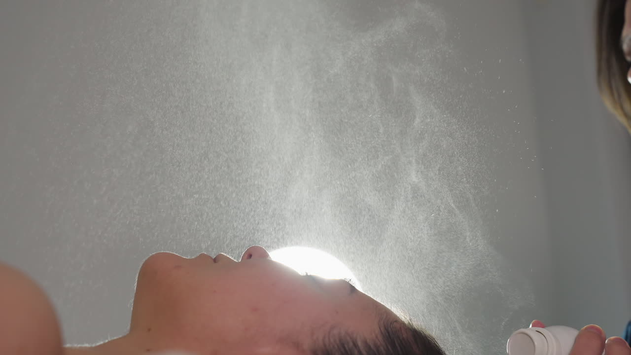 Professional hand spraying hydrating mist onto face of spa client under bright lamp for refreshing skin hydration and soothing relaxation during facial treatment session fine droplets