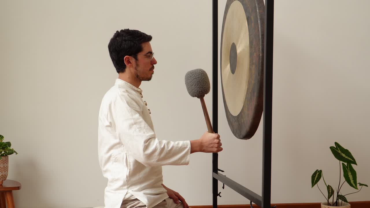 Profile view of a man kneeling on a cushion and striking a large symphonic gong with a soft mallet in a calm minimalist indoor studio with plants—ideal for meditation